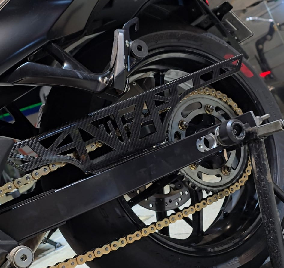 Close-up of a black motorcycle rear section with a gold chain, rear wheel, and a black metal chain guard in a garage setting.