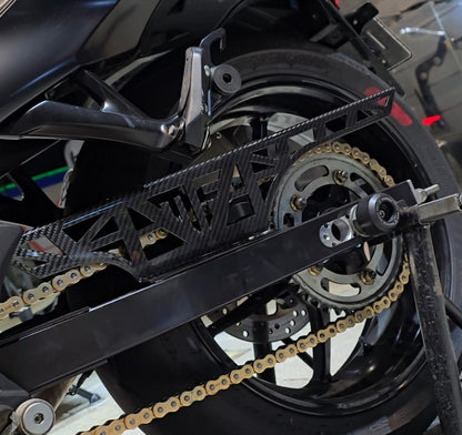 Close-up of a black motorcycle rear section with a gold chain, rear wheel, and a black metal chain guard in a garage setting.