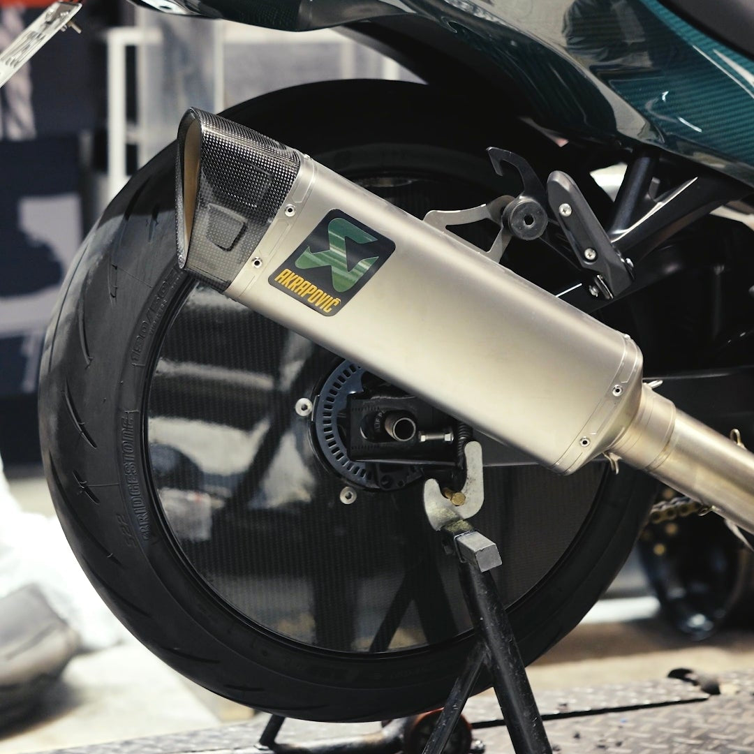 Close-up of a motorcycle rear section with a metallic silver exhaust muffler, black wheel, and teal-green body panel in a showroom setting.